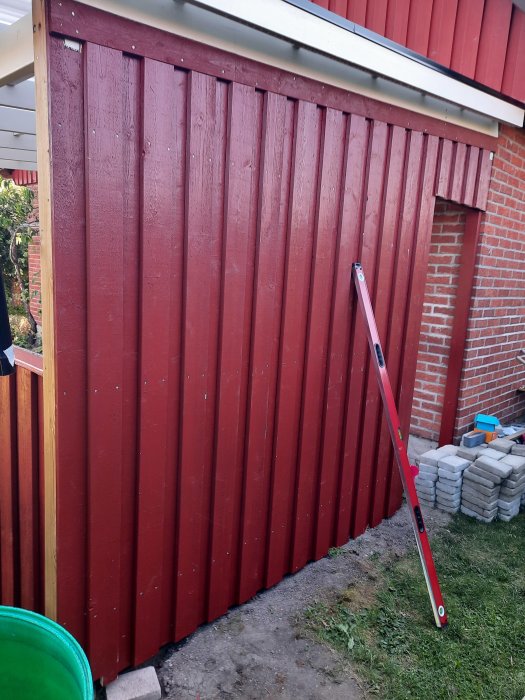 Nymålad röd träpanel på byggnad med vattenpass lutat mot den, bakom syns ett rött tegelhus.