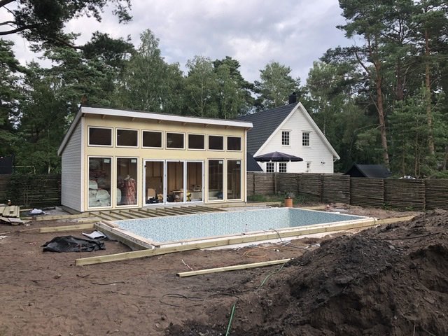 Byggarbetsplats med tömd pool och pågående trädäcksinstallation vid hus med skogsbakgrund.