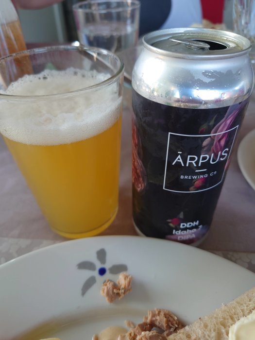 Ölglas fyllt med öl bredvid en öppnad ölburk av märket Årpus, på ett bord med matrester.