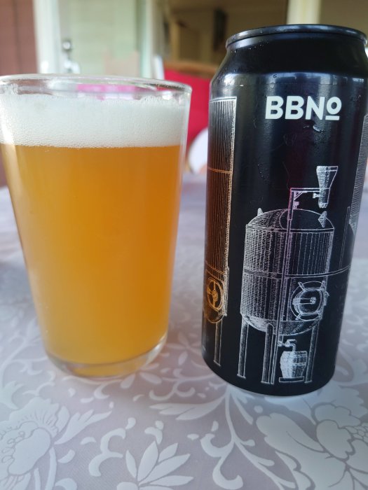 Ett glas öl bredvid en ölburk med etikett på ett blommönstrat bord.