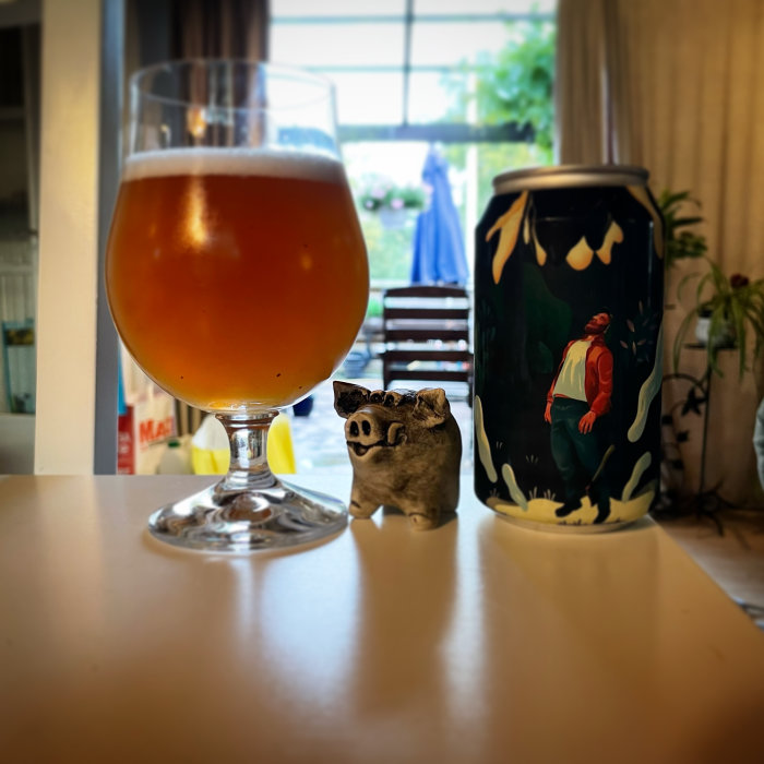 Ölglas med skummande öl framför en ölburk med konstnärlig design, liten grisskulptur på bordet.