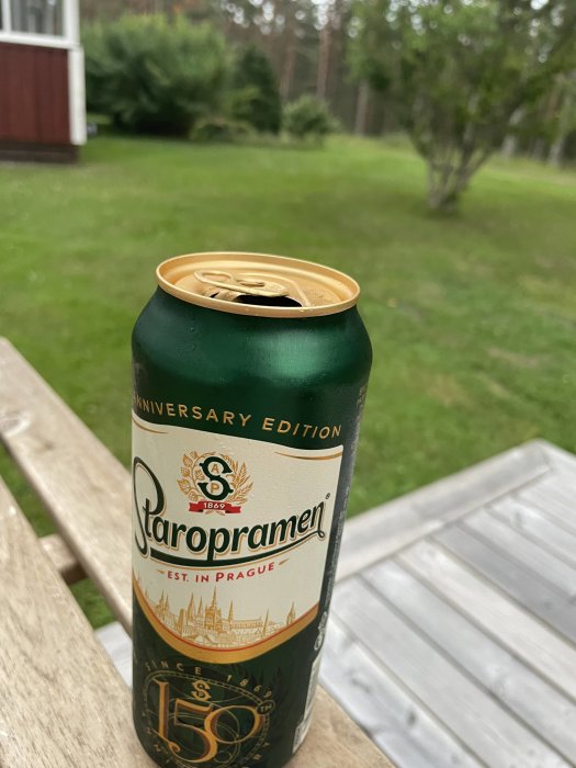 Öppnad Staropramen ölburk på ett utomhusbord med suddig trädgård i bakgrunden.