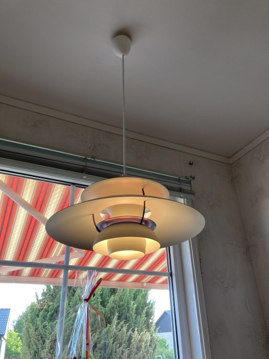 Retro taklampa med skärmar i nyanser av beige och rött, upphängd framför ett fönster.