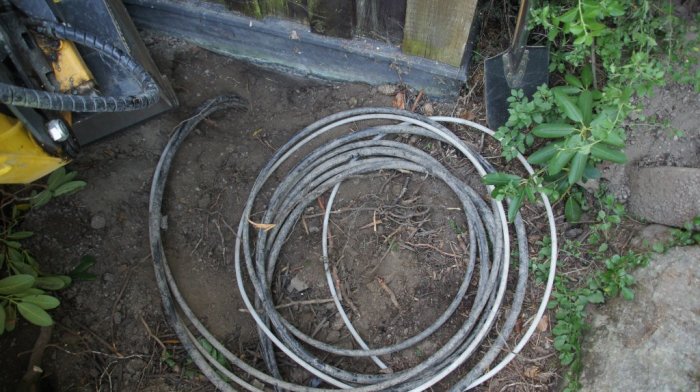 Knippe med tre övervuxna tunna slangar på marken bredvid grävverktyg och grönska.