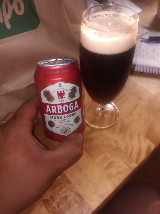 Hand håller en burk Arboga Mörk Lager med ett glas fylld med öl bredvid, på ett träbord.