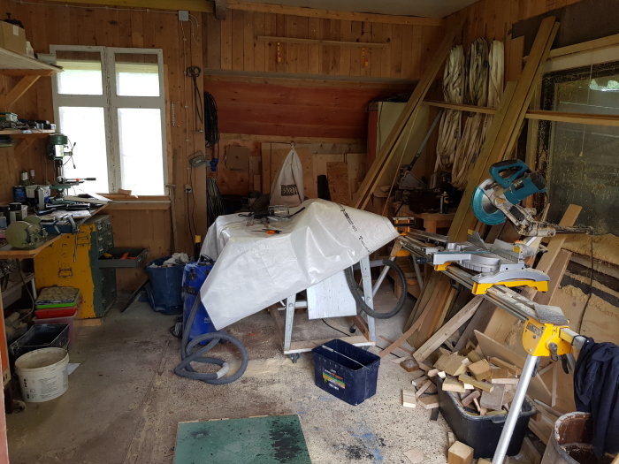 En rörig verkstad med träslöjdverktyg och material vid en bordssåg framför ett fönster med plywood.