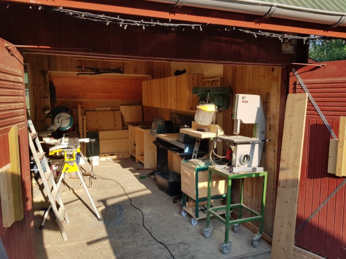 Garage med verktyg och maskiner, plywoodvägg där fönster satts igen, oorganiserad arbetsplats.