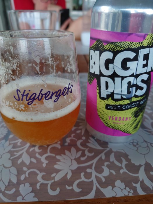 Halvtomt glas öl med Stigbergets logotyp och burk med texten "BIGGER PIGS WEST COAST VERDANT" på ett bord.