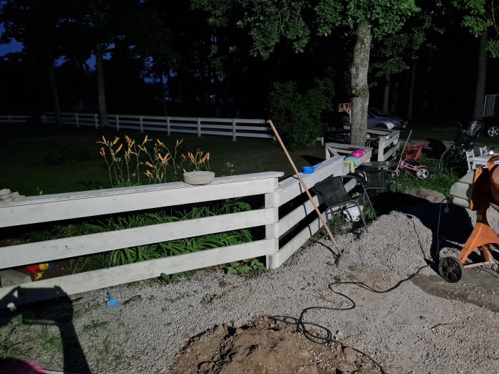 Trädgård på kvällen med öppen grind, staket, ek, leksaker och byggmaterial på marken.