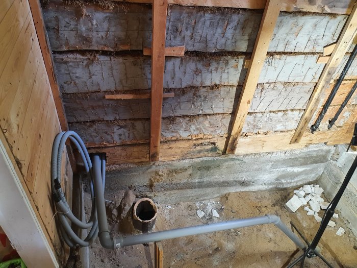 Bild av ett pågående badrumsrenoveringsprojekt med uppbrutet golv, synliga avloppsrör och råa timmerväggar.