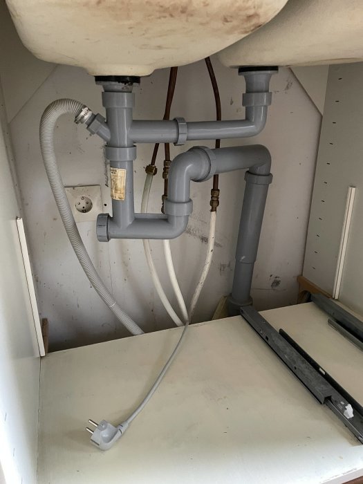 Vattenrör och avlopp under en diskbänk med osammanhängda delar och synliga elektriska kabel.