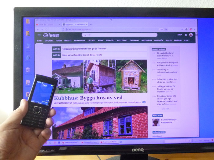Hand håller en äldre modell av en liten mobiltelefon framför en datorskärm som visar ett byggforum.