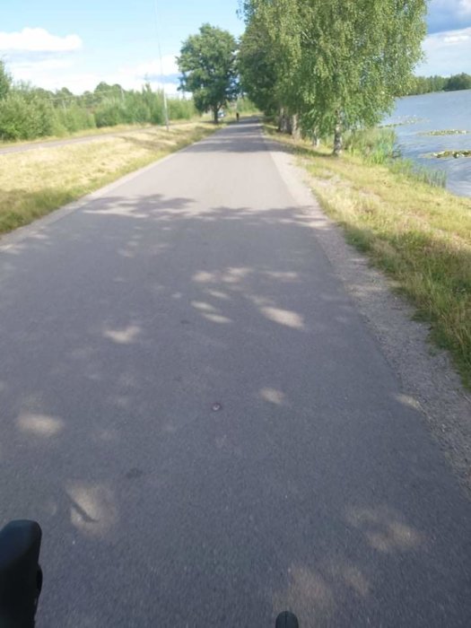 Asfalterad gång- och cykelväg längs med ett vattendrag omgivet av gröna träd och blå himmel, cykelstyre syns nederst.