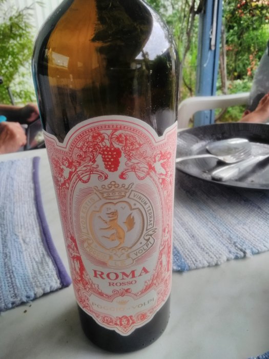 Flaska rött vin av märket Roma Rosso på ett bord, suddig bakgrund av en terrass.