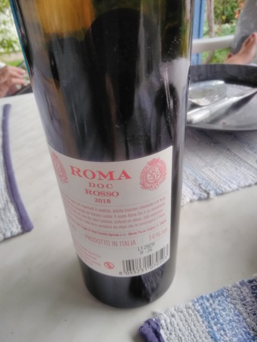 Flaska av Roma DOC Rosso 2018 rödvin på ett bord med en otydlig tallrik i bakgrunden.