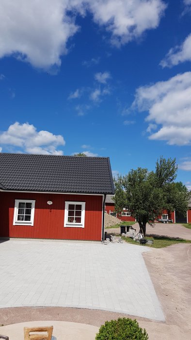 Ny lagd tumlad marksten i naturgrå och antracit framför röd stuga med blå himmel.