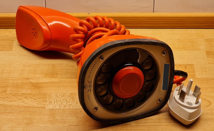 Dammig orange gammaldags telefon med nummerskiva och spiralkabel, liggande på ett träbord.