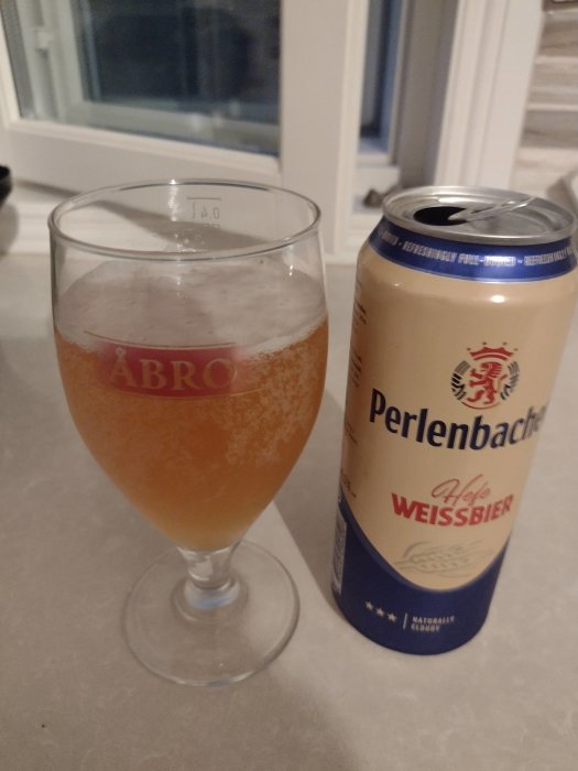 Ett glas med veteöl bredvid en öppnad Perlenbacher Hefe Weissbier burk på ett bord.