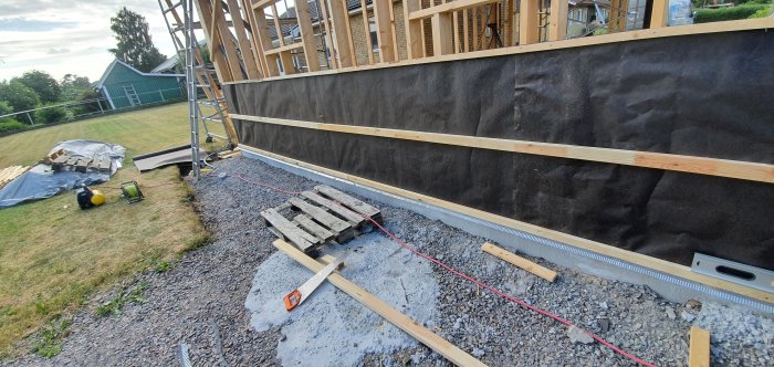 Nyinstallerad svart vindduk på trästomme med råspont vid husbygge, tillsammans med byggmaterial på marken.
