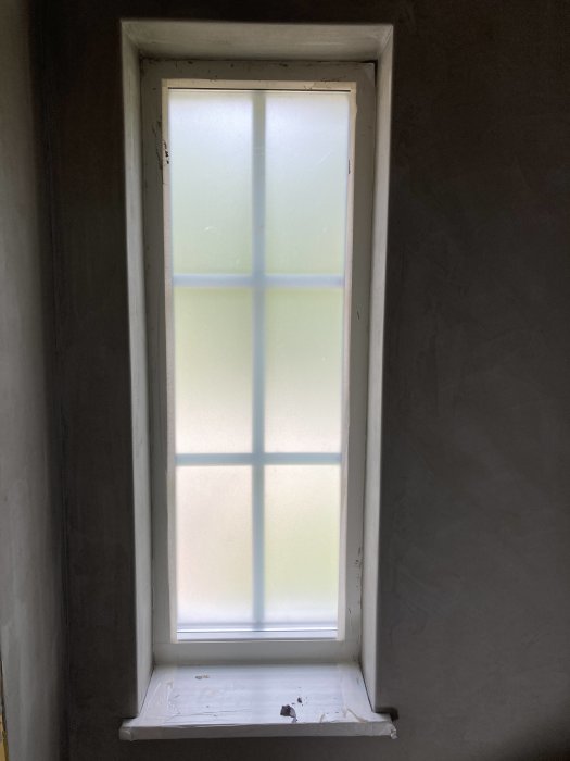 Vertikalt, smalt fönster med frostat glas och vita karmar inbäddat i en gråmålad vägg.