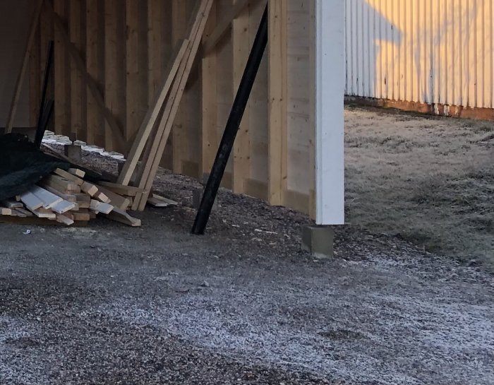 Carport på gjuten platta med trästolpar, gräsyta bredvid och synliga plintar under konstruktion.