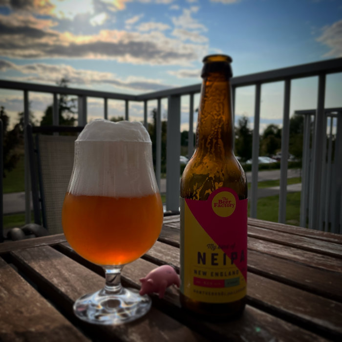Ölglas med skum på träbord vid solnedgång med en flaska NEIPA och en liten rosa plastgris.