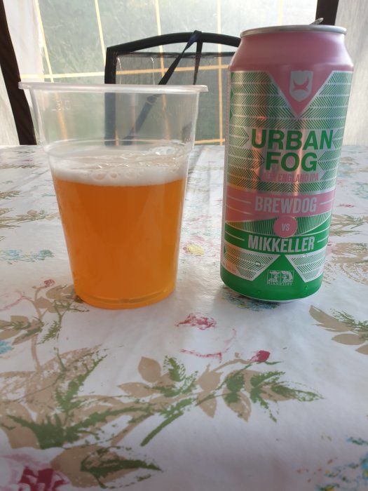 Burk av Urban Fog New England IPA av BrewDog vs Mikkeller bredvid ett plastglas med öl på ett blommigt bord.