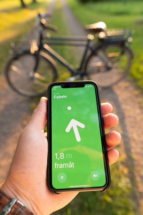 Hand håller en smartphone med en navigationsapp som visar "1,8 m framåt" med en suddig cykel i bakgrunden.