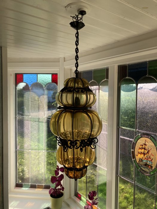 Hängande retro lampa med gyllene glaskupor framför ett färgglatt fönster.