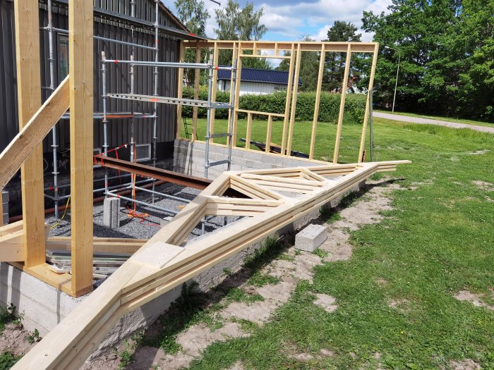Nybyggda trä-takstolar placerade på gräsmark intill en oavslutad byggkonstruktion och byggställningar under en klar dag.