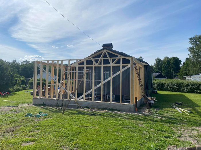 Hus under uppbyggnad med hemmagjorda takstolar och oskyddade träväggar på en gräsmatta.