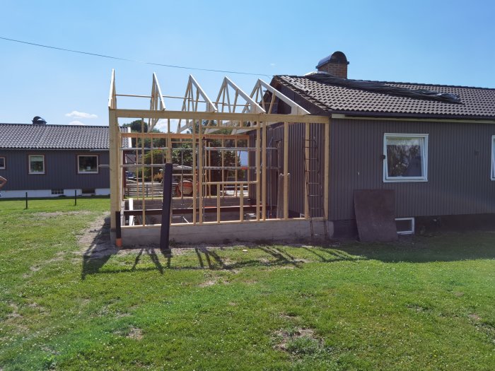Nybyggd trästomme med takstolar bredvid ett befintligt hus på en solig dag.