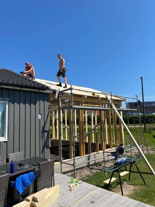 Två personer arbetar på ett hus med ofärdigt tak, verktyg och byggmaterial synliga på marken.