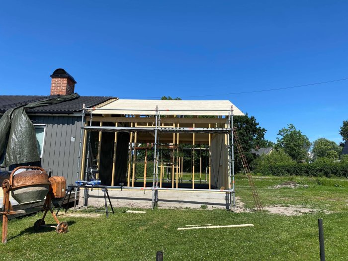 Husunderbyggnad med råspont på taket bredvid en befintlig byggnad under klar himmel.