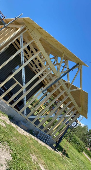 En stomme till en byggnad under konstruktion med synlig råspont och takstomme mot klarblå himmel.