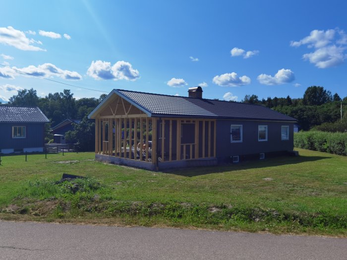 Hus under konstruktion med nytt tak och pågående arbete på en ofärdig veranda, beläget i ett soligt landskap.