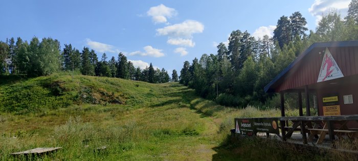 Naturscen med stig, grönt gräs, träd och en byggnad med skyltar och en röd sadeltak.