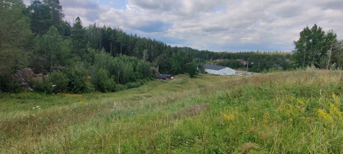 Landskap med en äng i förgrunden, en röd stuga bland träd i mitten och industrilokaler i bakgrunden, under en molnig himmel.
