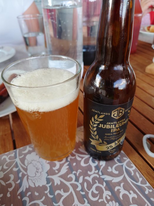 Ett glas med Jämtlands Jubileums IPA och flaskan bredvid på ett mönstrat bord.