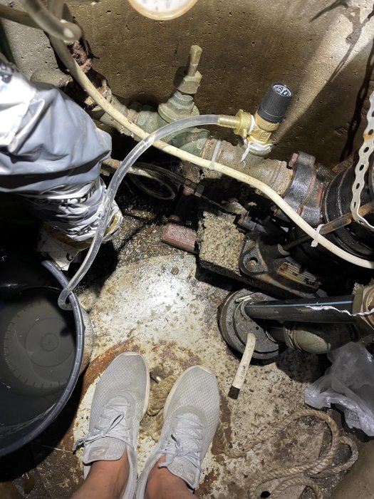 Fötter i sneakers bredvid en vattenpumpinstallation med slangar, ventiler och hink i ett vått och smutsigt utrymme.