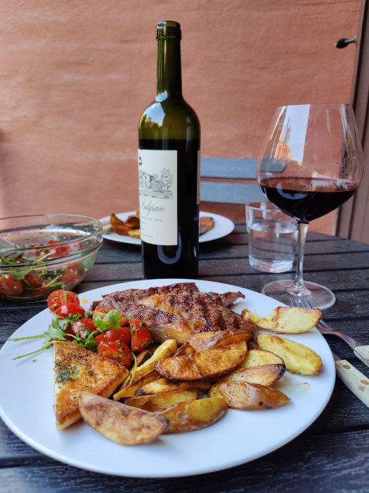 Grillad ryggbiff med kryddad stekost, rostad potatis, tomat, persilja på tallrik med vinflaska och glas.