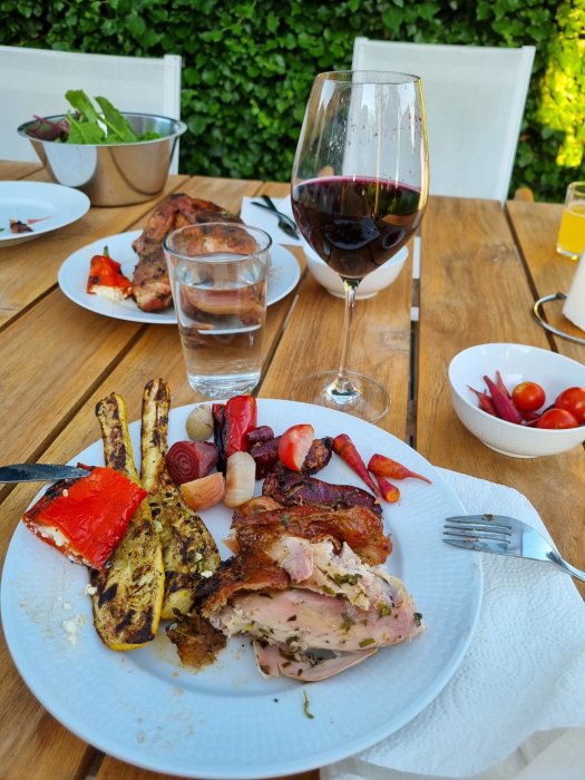 Grillad kyckling på tallrik med rotgrönsaker, ett glas rött vin och sallad på ett somrigt trädgårdsbord.