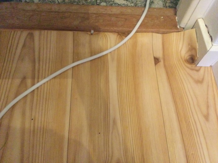 Golv med trästruktur syns delvis under grön plastmatta vid en väggkant med kabel på golvet.