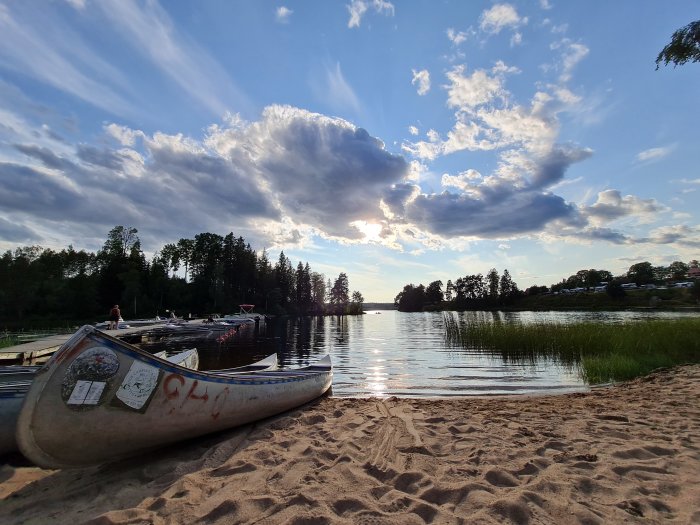 Kanot på strandkanten med sjövy, molnig himmel och solnedgång i bakgrunden.