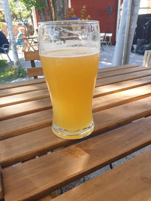 Ett glas med grumlig NEIPA-öl på ett träbord, utomhus med bakgrund av en uteservering.