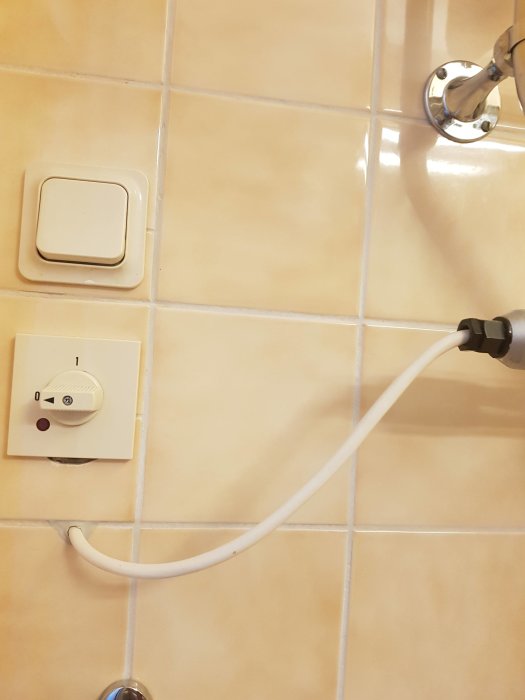 Alt: Vägg i badrum med ljusgula kakelplattor, eluttag och draget elsladd till en handdukstork.