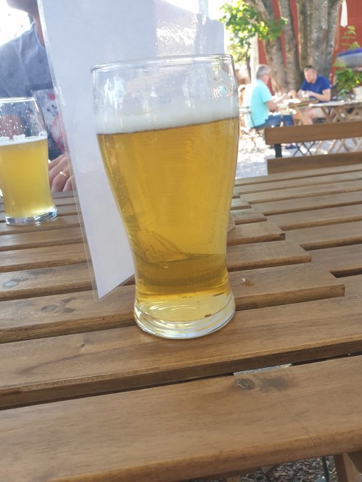 Ett glas Leksands pilsner på ett träbord utomhus med människor i bakgrunden.