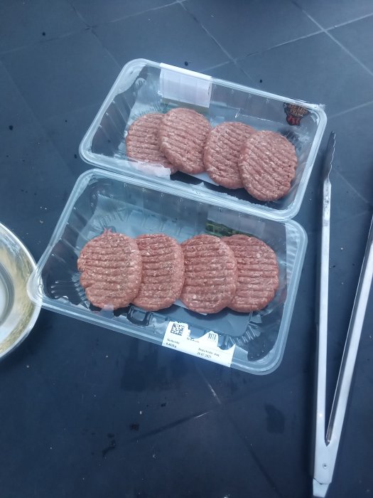 Förpackade köttfärsbiffar på ett bord bredvid en grilltång och en skål med tsatsiki.