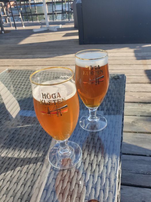 Två ölglas på ett utomhusbord vid en marina, med texten "HÖGA KUSTEN" på glasen.