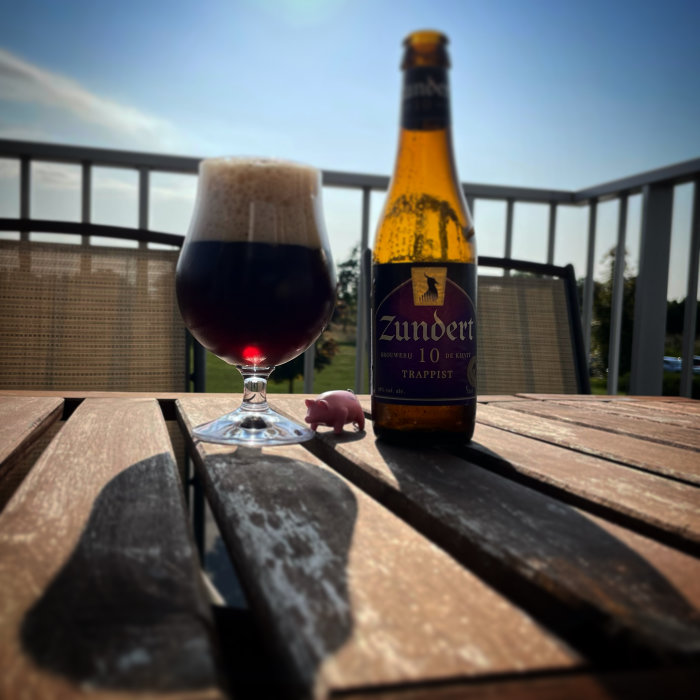 Ett glas mörkt öl och en flaska Zundert Trappist på ett träbord utomhus i solskenet